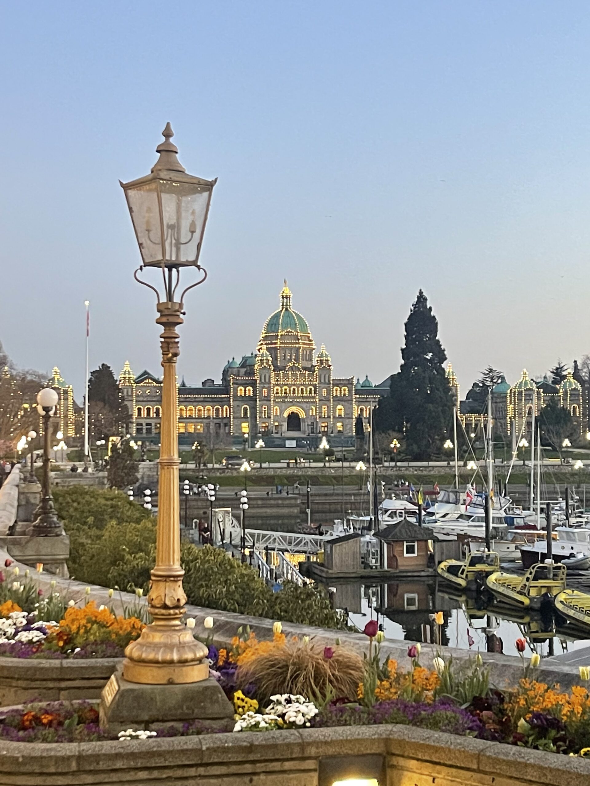 View of Victoria Inner Harbor during sunset looking towards the lit up Legislative Administrative building with a beautiful street lantern in the foreground. In front of the building are moored boats
