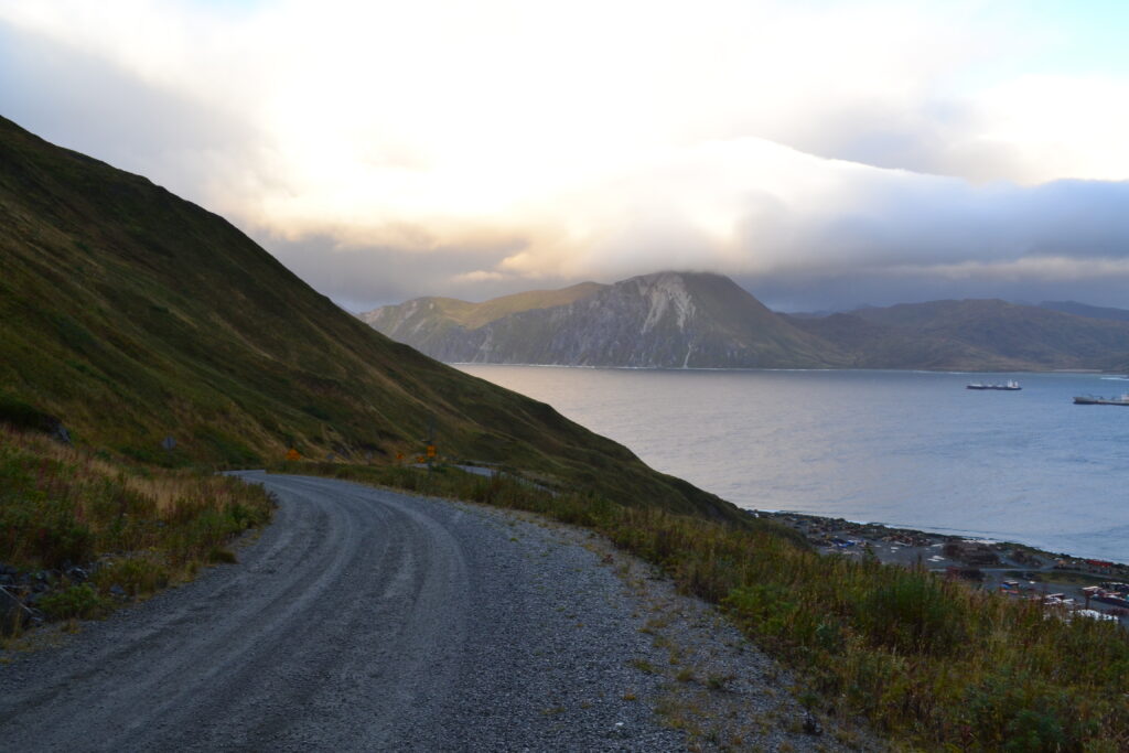 Road going down a mountain with a bay that has two boats and a mountain in the backdrop