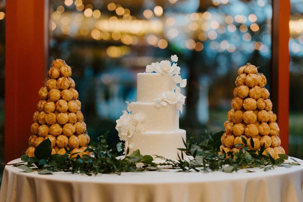 Two towers of cream puffs on either side of a three tiered white wedding cake on a table with greenery