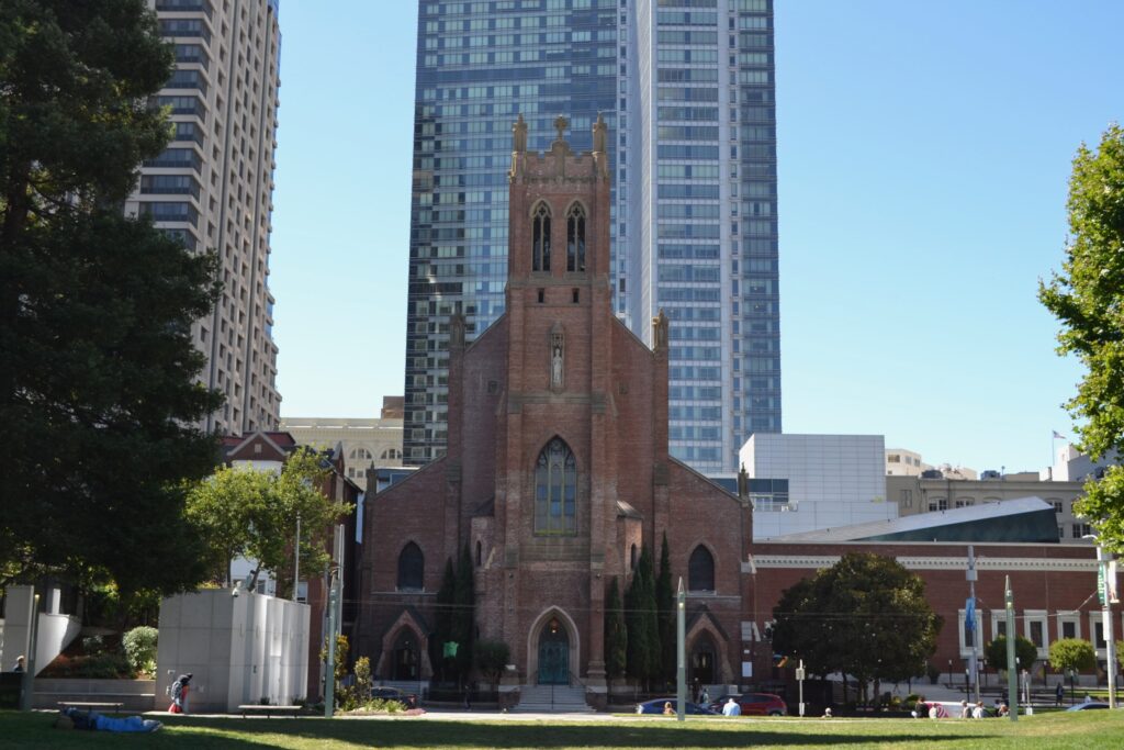 Brick Catholic church with bell tower in the middle stands in front of a glass high rise in San Francsico