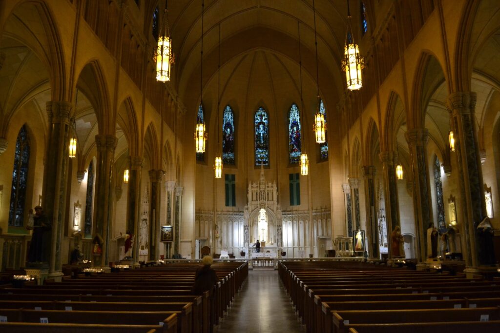 The nave of a Catholic Church with pews and columns going down both sides and a High altar at the front surrounded by stained glass windows.
