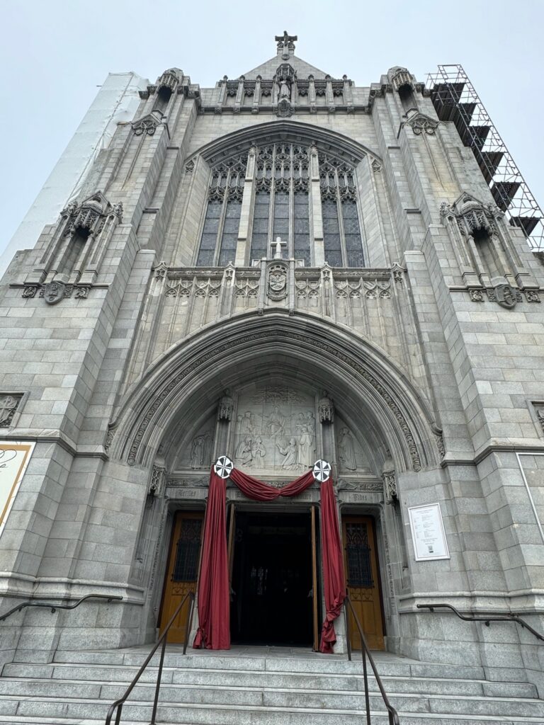 The facade of a gothic church, the arms of the Dominican order hold a red draped curtain above the entrance. Statues, stained glass and scaffolding can be seen along the church