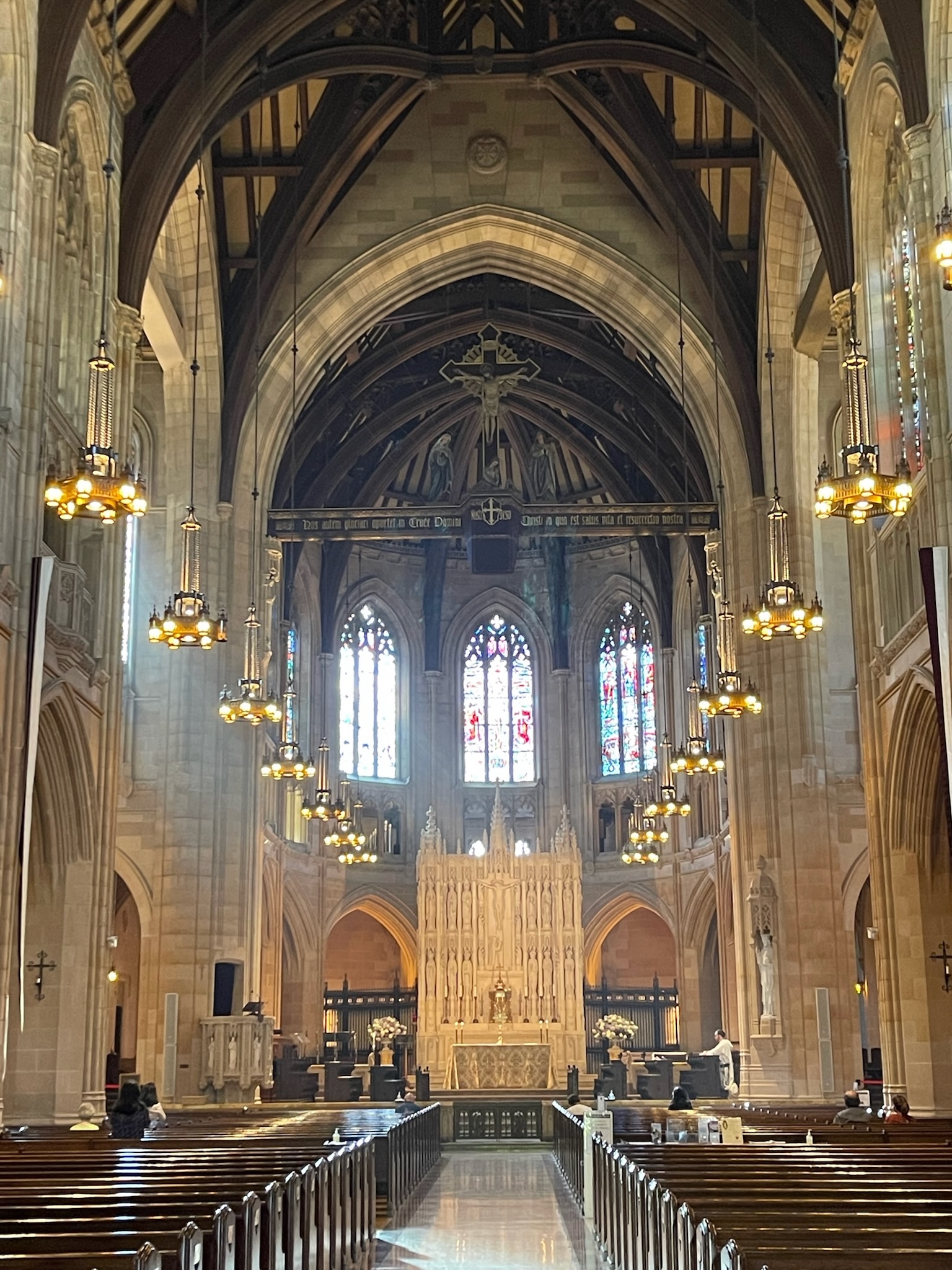 A nave of a gothic church, pews in the foreground, lead to a high altar and stained glass windows in the background.