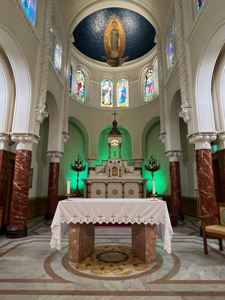 Altar of a Catholic Church with high altar behind it illuminated by green lights. Domed ceiling above with mural of Virgin Mary and stained glass windows