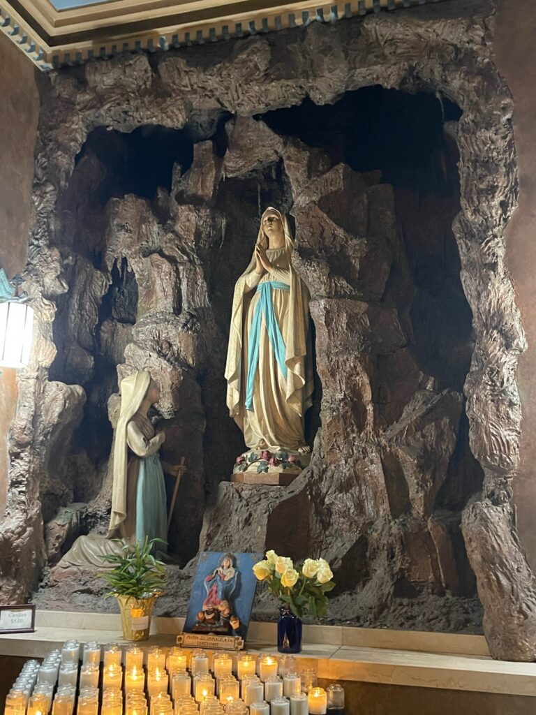 Rows of candles, two bouquests and an image of Mary in the foreground lead to two religious statues inside imitation rock.