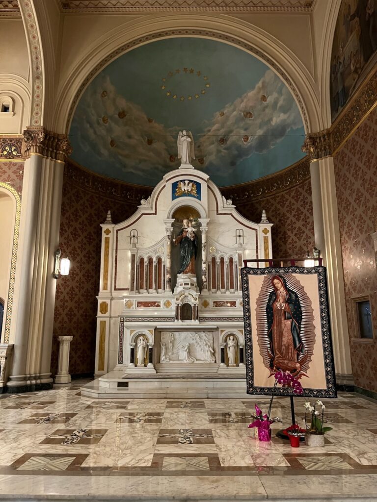 A marble altar with beautiful statues, above a fresco of heaven. In the foreground is an image of Our Lady of Guadalupe