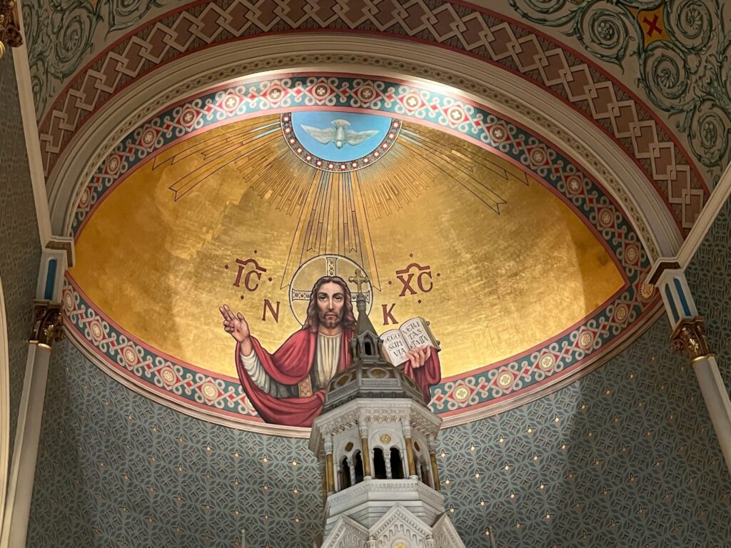 A fresco of Jesus Christ with the Holy Spirit dove above in a Golden Background form the interior of a half dome.