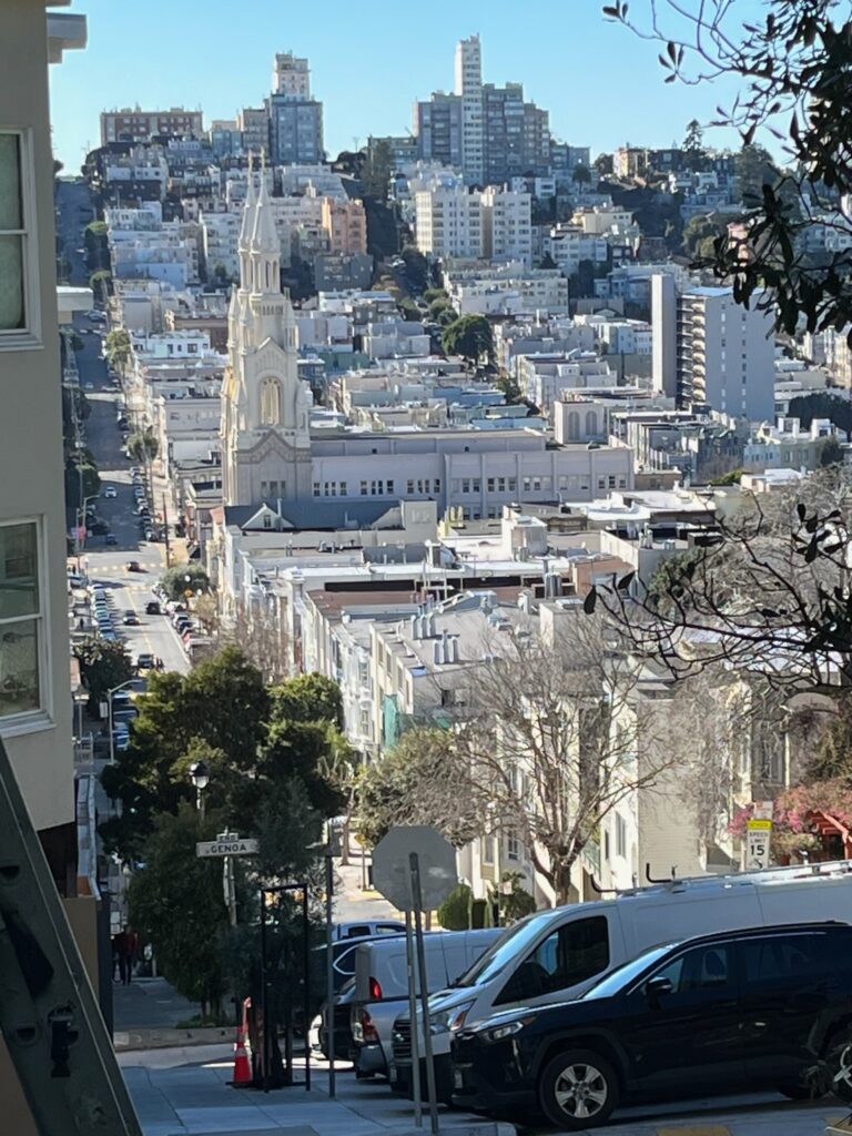 A street with parked cars slopes down an urban hill to a large church, and slopes up again with buildings built all around it.