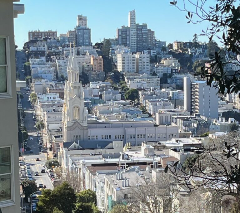 A street with parked cars slopes down an urban hill to a large church, and slopes up again with buildings built all around it.