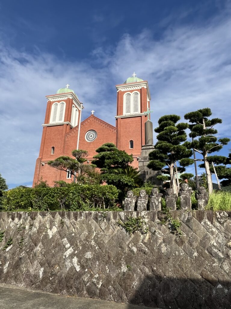 A stone wall, with statues and trees, behind a brick catholic church with two spires