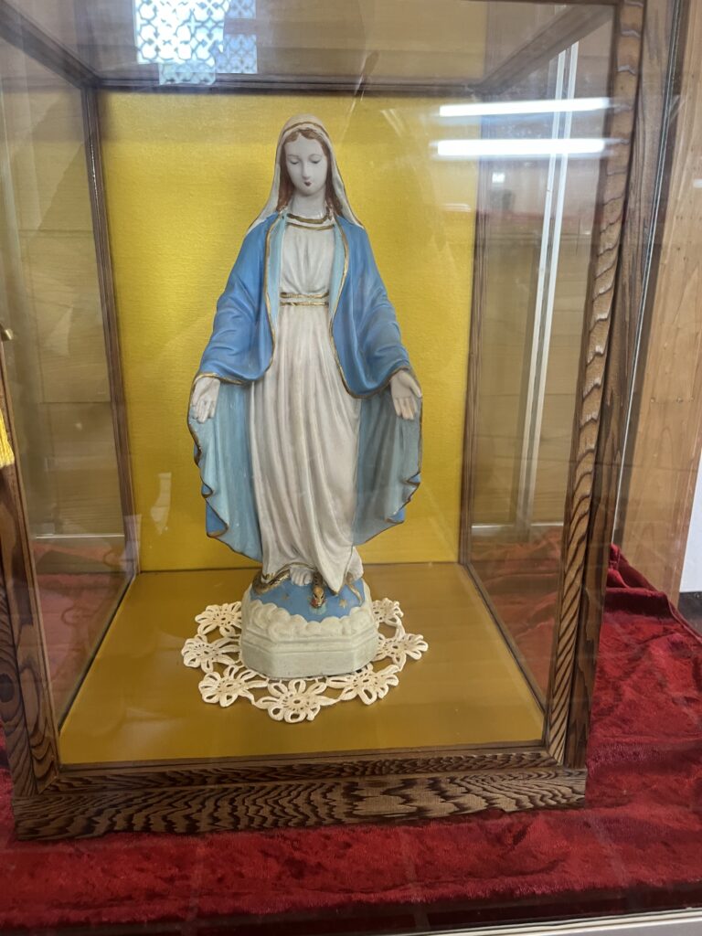 A statue of Mary with her arms out inside a museum display, with a yellow background