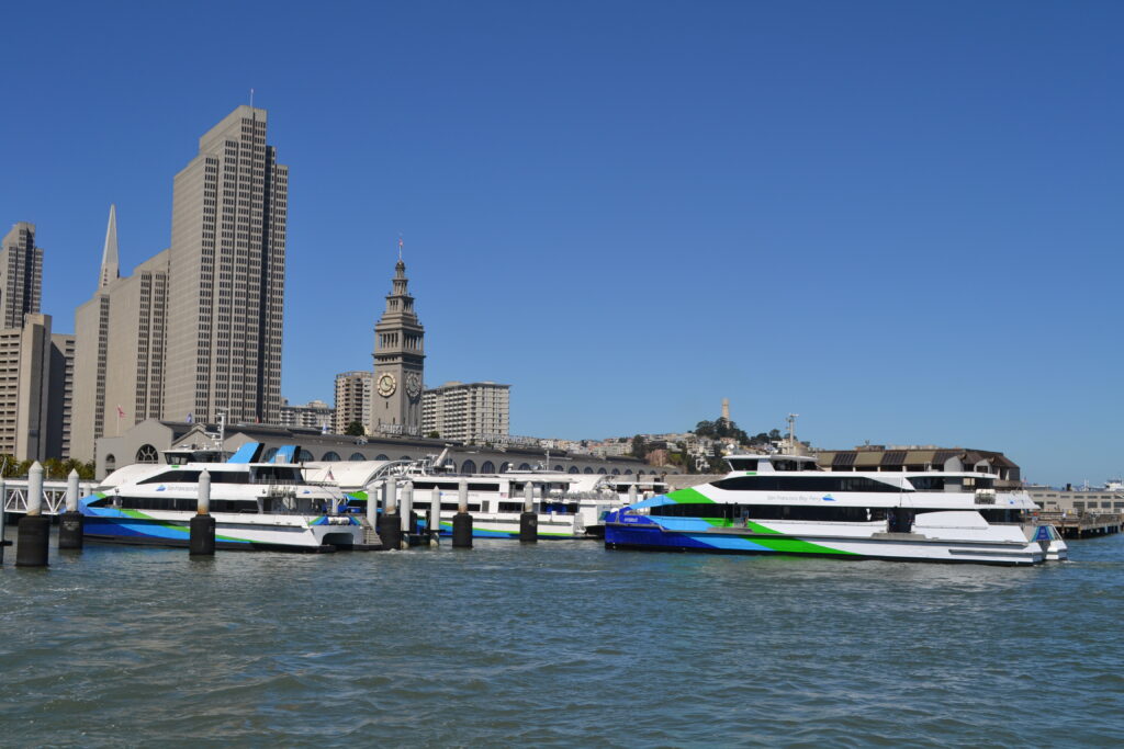 A ferry approaches a pier with high rises, a ferry building and a tower on a hill in the background