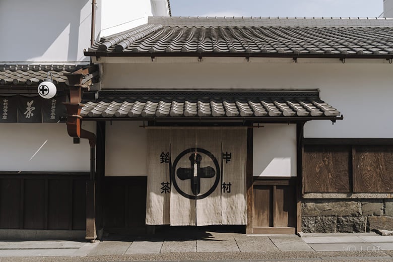 The traditional wooden exterior of the Nakamura Tokichi tea shop in Uji, Japan, featuring a white noren curtain with a black circular logo and grey tiled roofs.