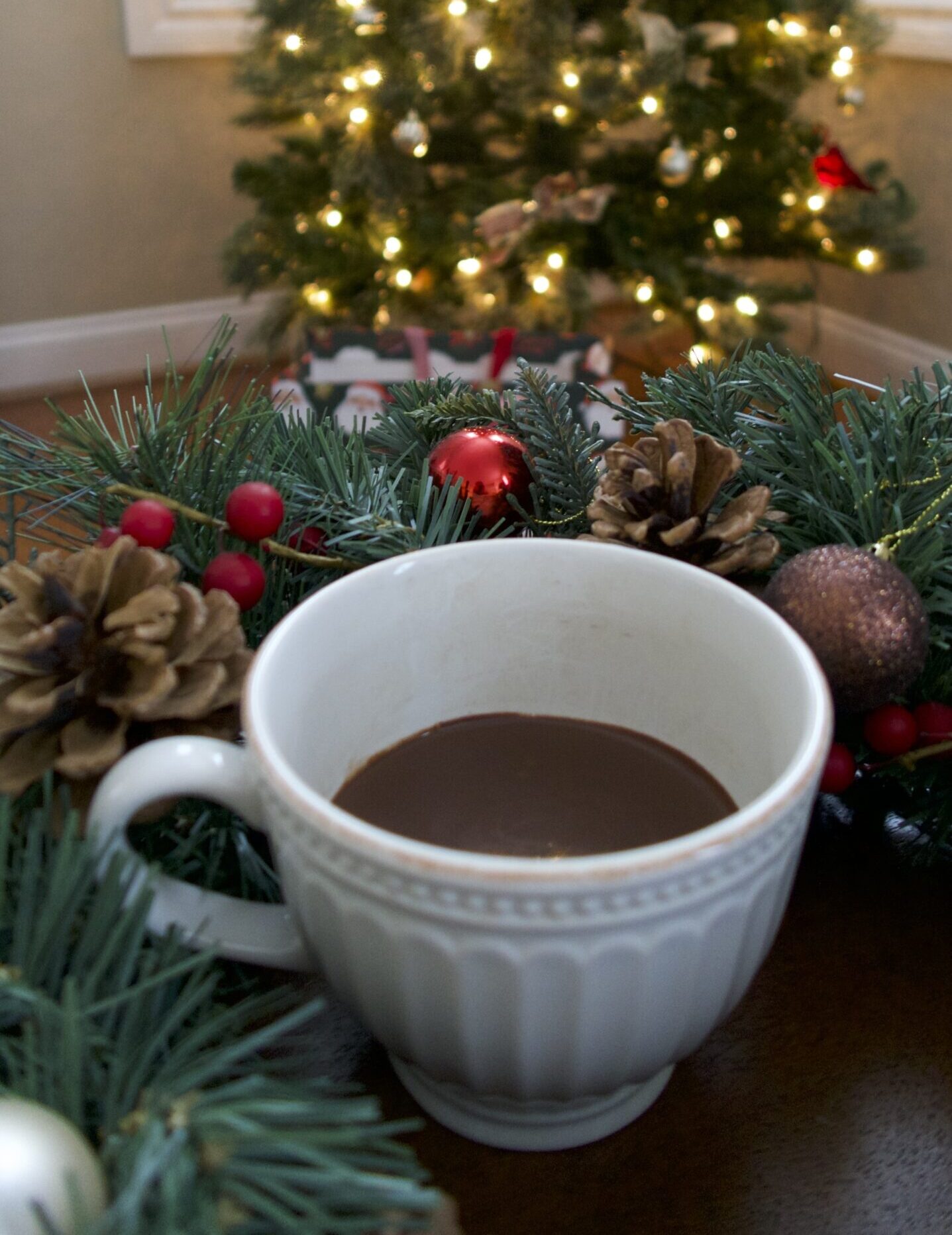 mug of hot chocolate surrounded by garland and in front of a Christmas tree