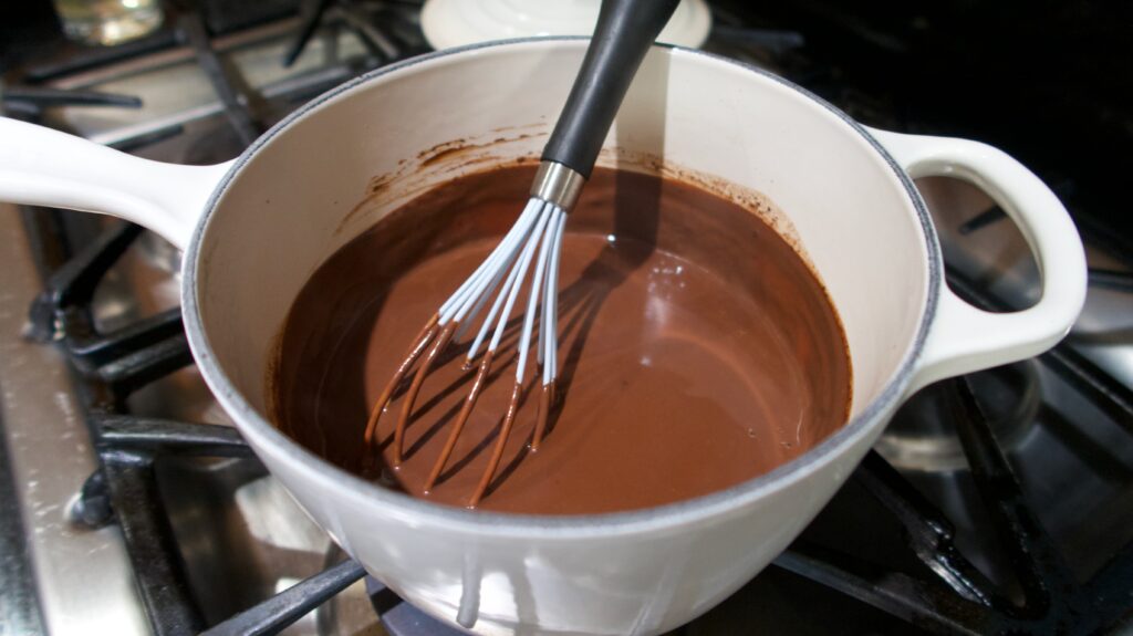 a saucepan on a stove with thick hot chocolate inside being stirred by a whisk