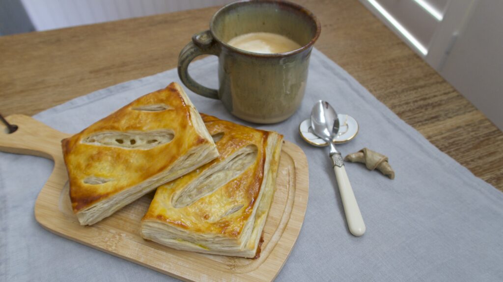 Puff pastries on a wooden cutting board next to a cup of coffee and some utensils on a blue linen napkin