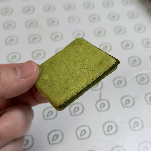 A close-up of a Matcha Chitose Langue de Chat cookie being held. The thin, bright green matcha-infused square wafers sandwich a thick layer of dark green matcha chocolate filling.