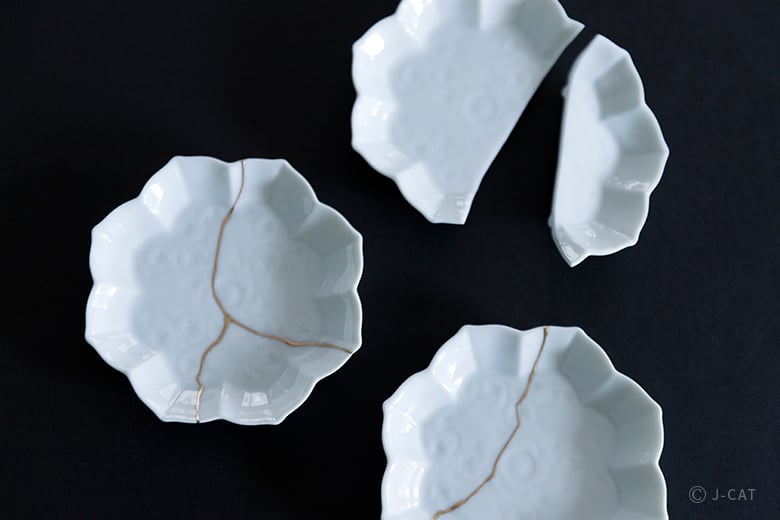 Three white ceramic flower-shaped plates on a black background, showcasing the Japanese art of Kintsugi with gold-lacquered seams joining broken pieces.