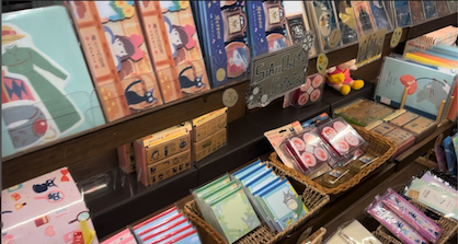 A wide variety of official Studio Ghibli stationery products arranged on store shelves. The collection includes illustrated notebooks, small memo pads in wicker baskets, and themed packaging featuring iconic characters like Totoro and Kiki.