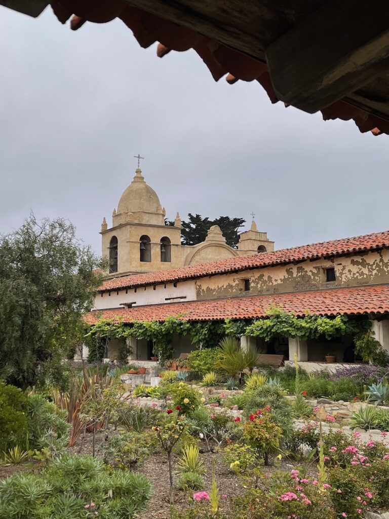 Carmel Mission building in the background with a garden in front that has landscaping with all colors of flowers