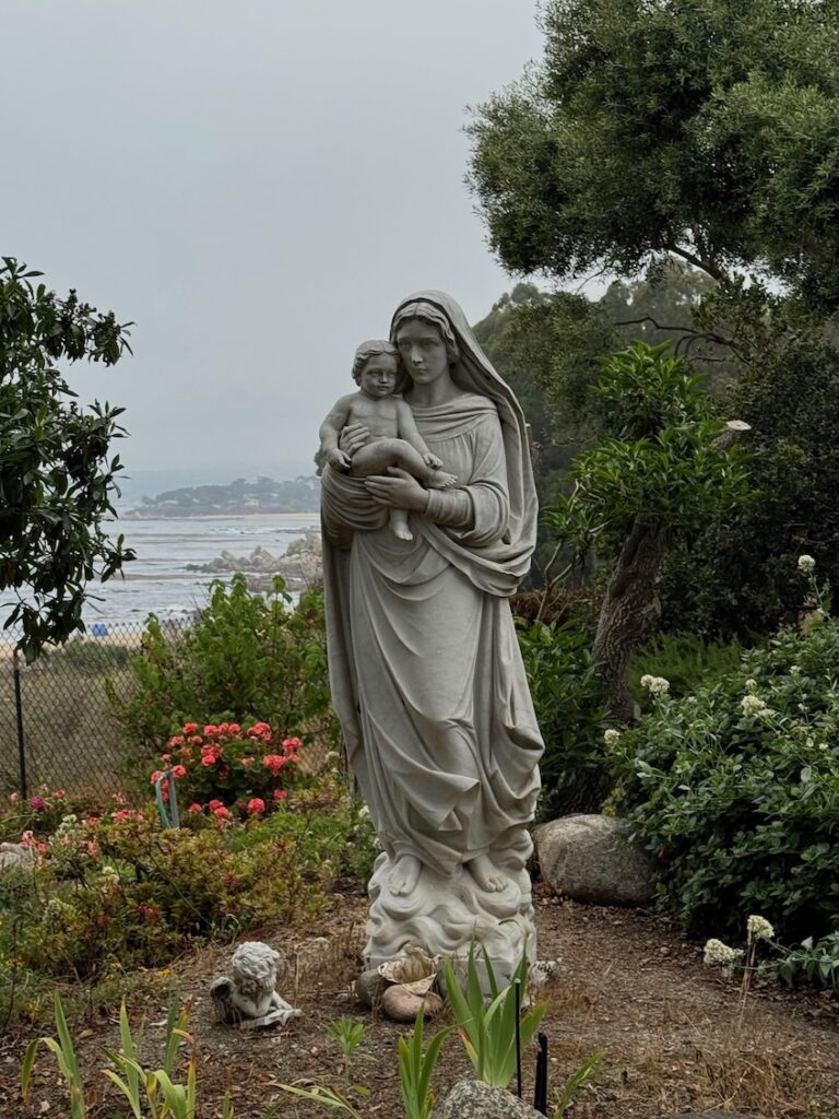 Life-sized stone statue of of Virgin Mary holding Baby Jesus in a garden with an ocean view in the background