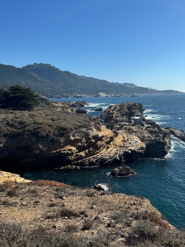 Rocky cliffs meet the ocean in Northern California on a clear day with rolling hills in the background