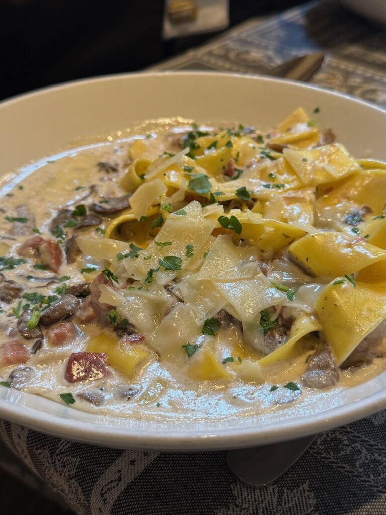 Plate of carbonara with parpadelle pasta and bits of pancetta, basil, and shaved parmesan shaved on top