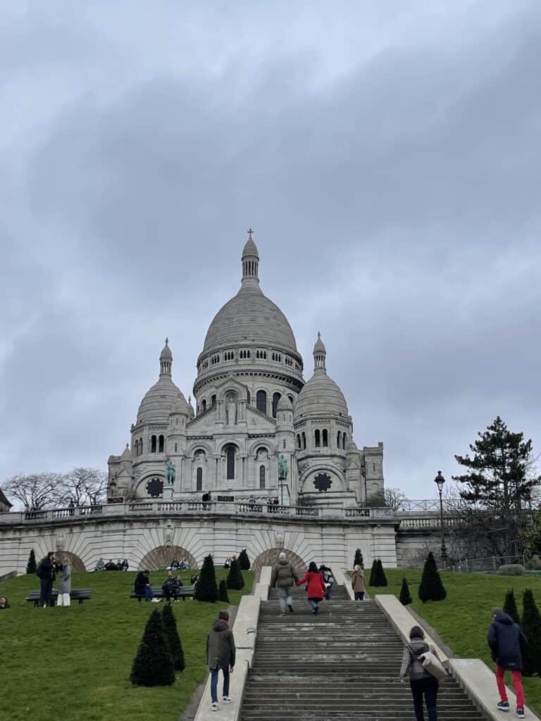 Majestic Sacré-Cœur Basilica in Montmartre, Paris, featuring its iconic white domes against a cloudy sky, surrounded by visitors exploring the historic site and scenic gardens.