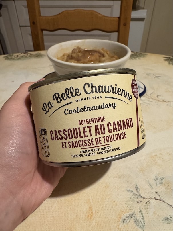 A hand holding a can of cassoulet with a bowl of cassoulet in the background on a table
