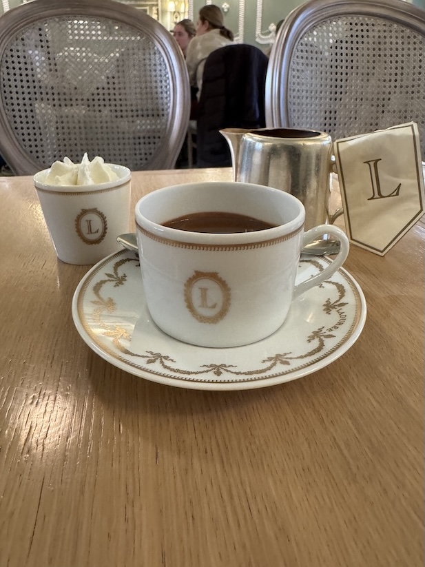 A mug of hot chocolate on a little plate with a small pitcher in the back and a cup of whipped cream