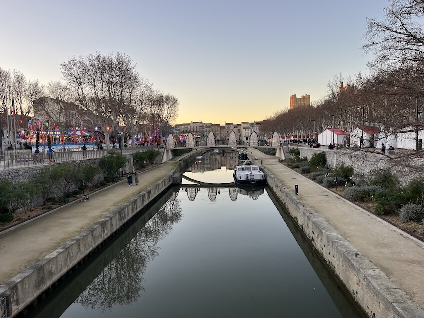 French canal with a bridge over it has an amusement park on the left side and a Christmas market with stalls on the right side
