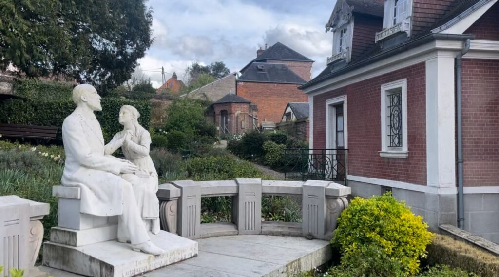 Statue of St. Therese pleading with her father in a garden behind a brick house