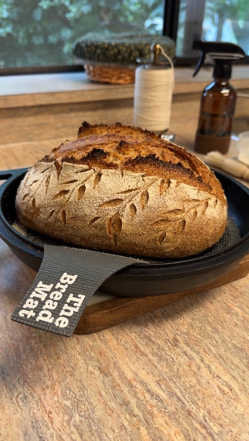 A whole wheat sourdough boule with wheat stalk designs on it is on top a cast iron cooker on a counter with a twine stool and spray bottle in the background
