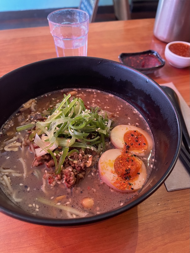 A steaming bowl of ramen topped with sliced green onions and savory ground pork, nestled in a rich broth with noodles visible beneath. The vibrant green onions add freshness and color contrast to the hearty, flavorful pork and warm soup. A halved soft-boiled ramen egg with a slightly runny yolk floats on top