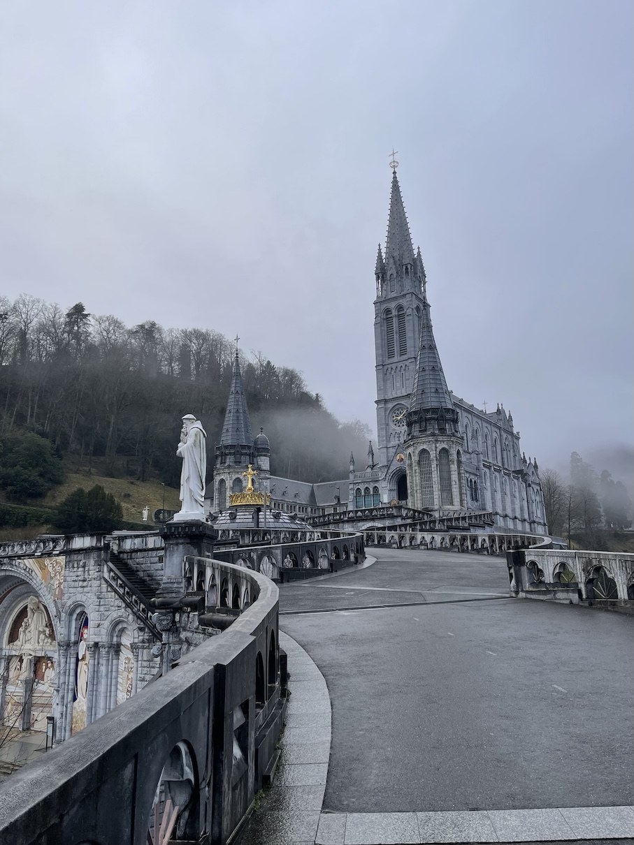 The path leading up to the Basilica of the Immaculate Conception in Lourdes, France which is the upper level basilica with a foggy forest in the background and a statue along the railings leading up to the basilica