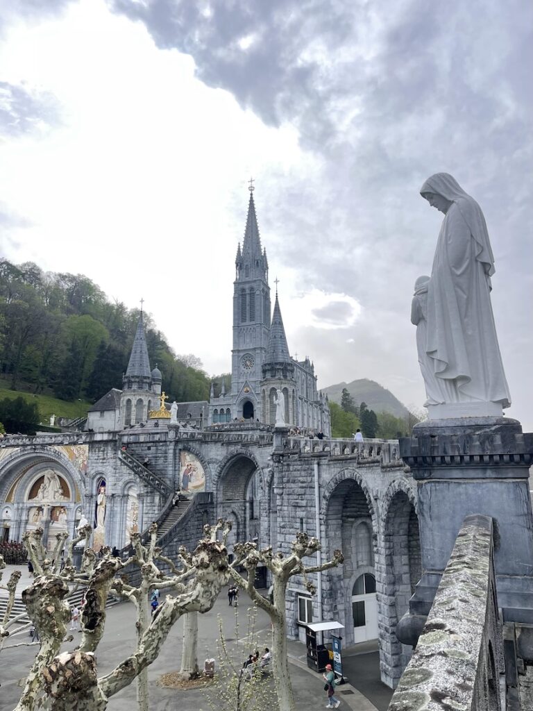 Picture of the basilica at the sanctuary of Lourdes taken from the side path leading up to the upper basilica. There's a statue of St. Anne on the side toward the front of the frame