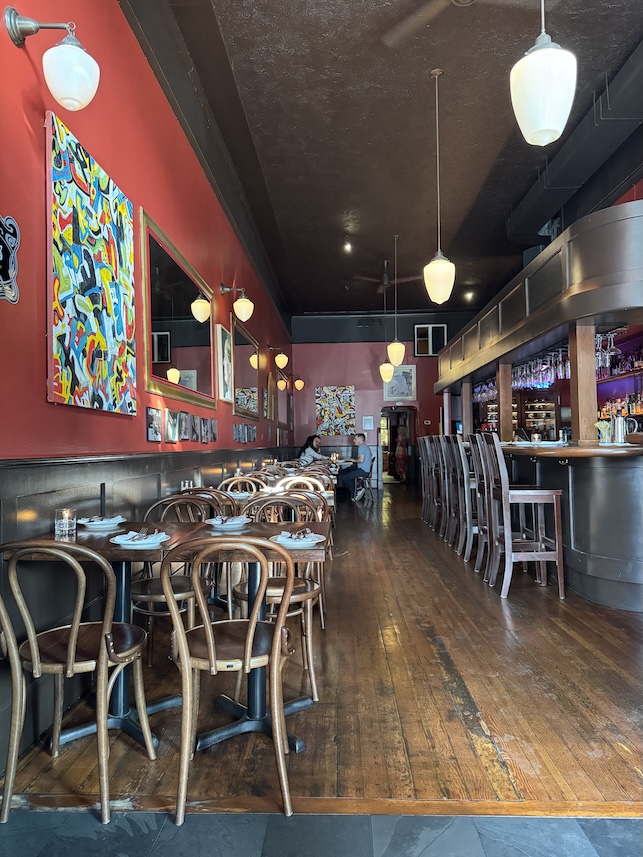 The interior of a restaurant with tables on the left and a bar on the right, the walls are painted red with modern art displayed. Ceilings are painted a dark brown