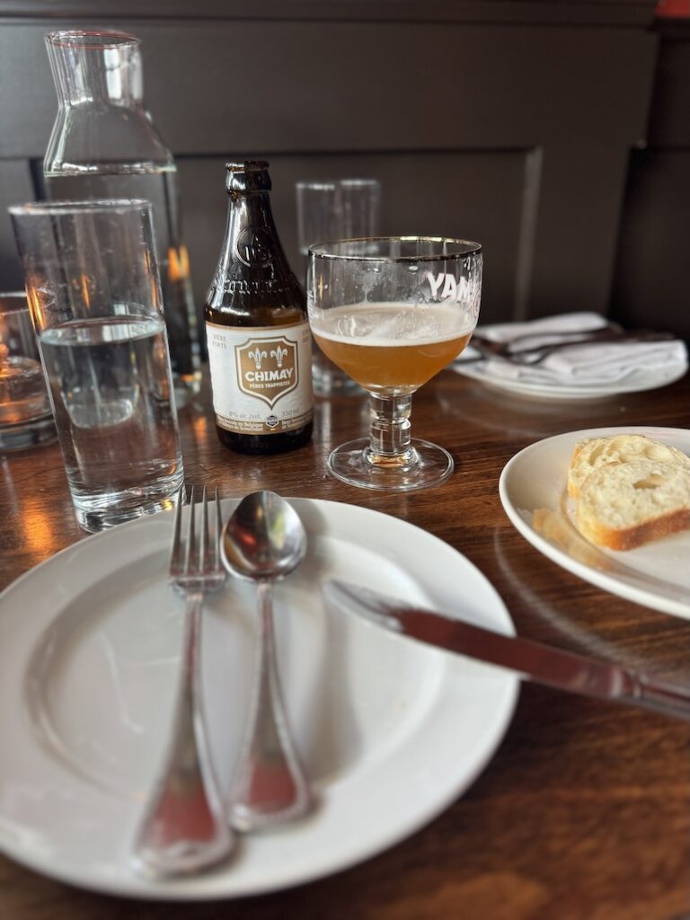 A tabletop at a French restaurant with silverware arranged on the plates and a chimay bottle of beer poured into a gauntlet