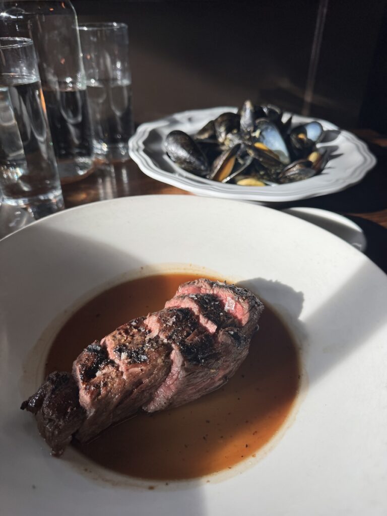 A wooden table with two dinner plates on top with the one in the front having a steak sliced sitting in sauce and the back plate contains steamed mussels