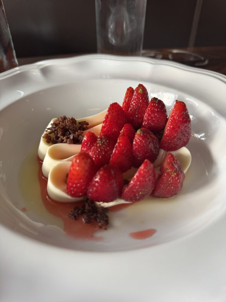 A dessert plate of white chocolate ganache piped beautifully with strawberries cut in half arranged neatly on top. Sprinkled over it are also toffee bits all over a strawberry syrup on the bottom
