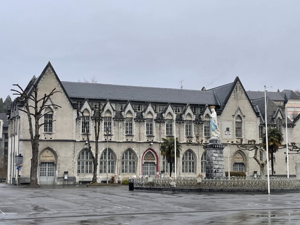 A beautiful building dedicated to the sacrament of Reconciliation in the Sanctuary of Lourdes, France with two stories and many arched windows across the building. There is the statue of Our Lady of Lourdes in front of the building at the center of the Sanctuary