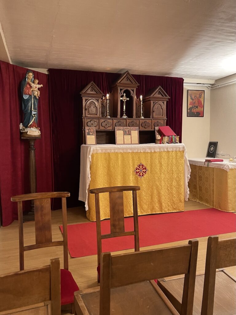 Small chapel with wooden high altar that has a crucifix in the center with an altar below covered in a gold fabric. There are red velvet curtains all along the wall and chairs for pews