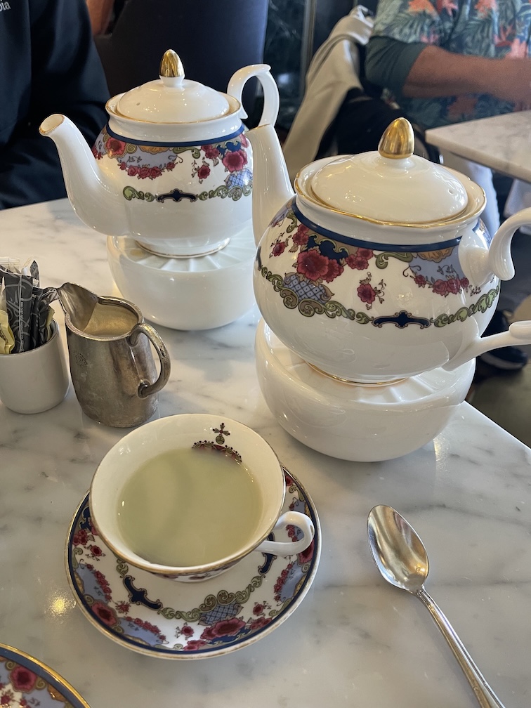 On a white marble table are two fancy china teapots sitting on teapot warmers as well as a teacup on a saucer with green tea in it that is opaque with a bit of cream. On the table is also a silver teaspoon and silver pitcher for the cream