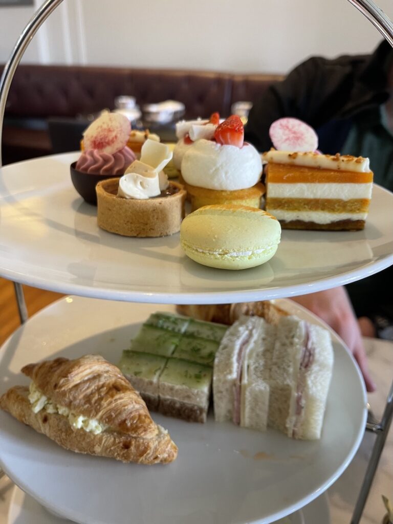 An afternoon tea stand with two levels containing sweet tarts and desserts on the top followed by sandwiches and croissants on the tier below.