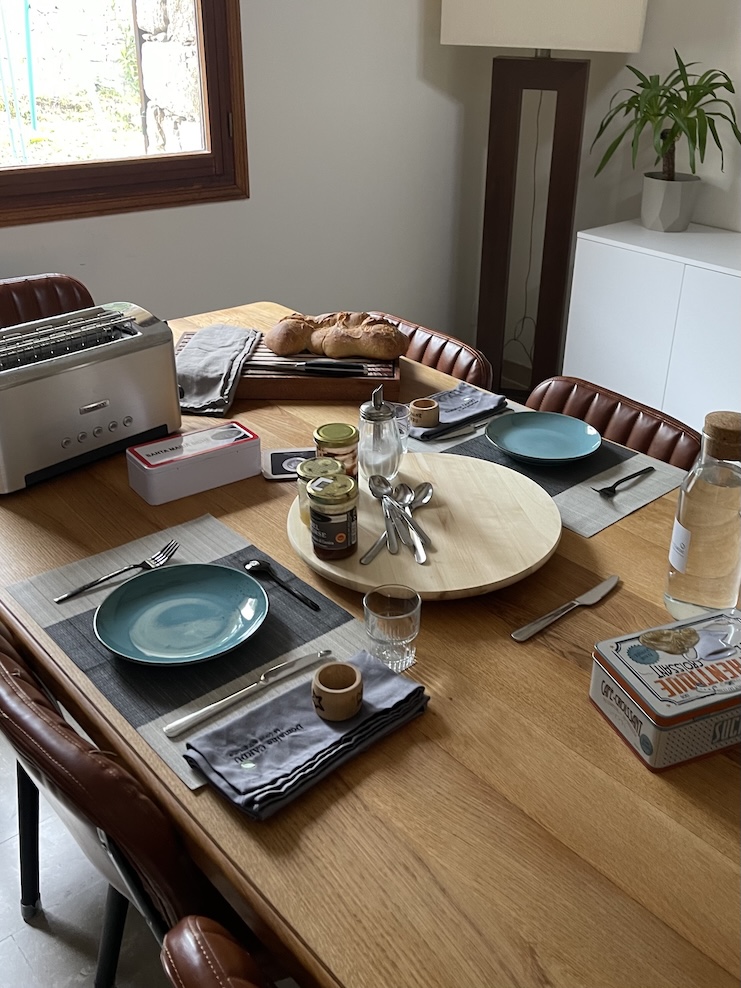 A wooden dining table set for breakfast with teal plates, cutlery, and glasses on placemats. In the center is a round wooden tray holding jars of spreads, with fresh bread on a cutting board nearby. A toaster, bottled water, and packaged biscuits are also on the table.