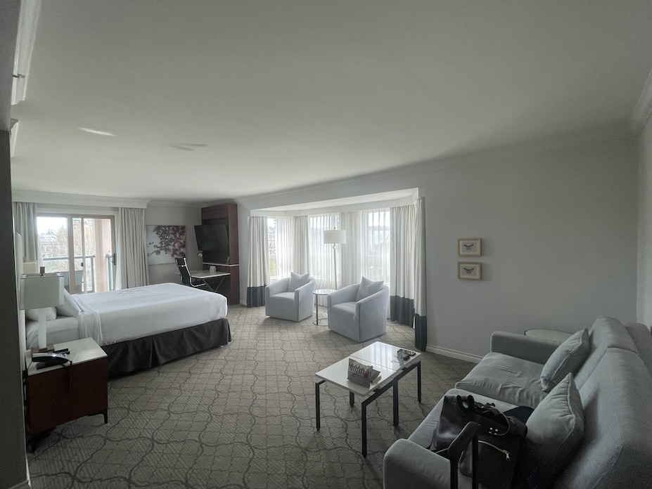 A cozy and elegant king bed room at the Hotel Grand Pacific in Victoria, BC. The room features a large, neatly made king-sized bed with plush white linens and pillows. gray, neutral tones and soft lighting create a relaxing atmosphere. A wooden nightstand with a lamp sits beside the bed, and two comfortable armchairs are positioned near a window that offers natural light. Along the wall is a large gray loveseat. Tasteful artwork and classic desk and tv set up complete the inviting space.