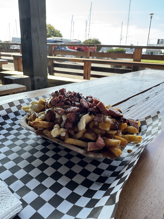 A poutine dish with smoked meat, cheese curds, gravy, and fries on a checked mat on top of a wooden table. In the background is a parking lot with boat masts in the back