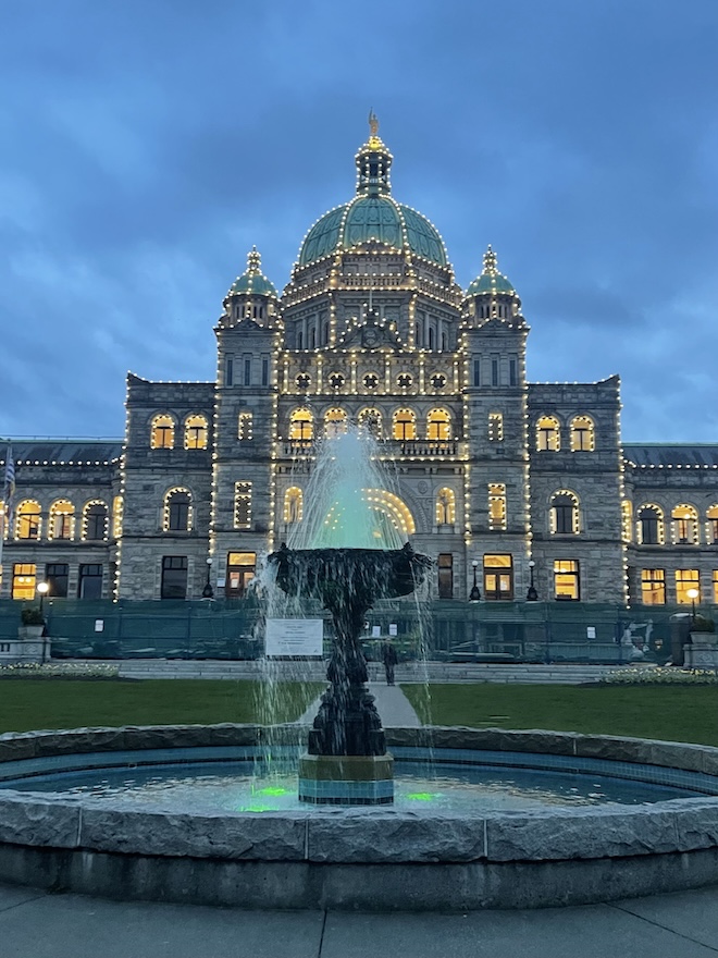 he British Columbia Legislative Building in Victoria at dusk, illuminated by warm lights that highlight its grand architecture. The building stands majestically behind a gently flowing fountain in the foreground, whose water reflects a glowing green light. The sky is a deepening twilight blue, adding a serene and dignified atmosphere to the historic scene.