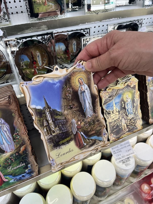 A hand holding a picture of Our Lady of Lourdes and St. Bernadette in a souvenir shop with other pictures on in the background and candles on the shelf below