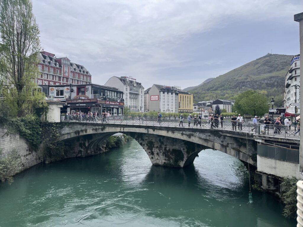 A small bridge over a blue-green river in Lourdes, France with pedistrians walking across and hotels in the background and a hill on the right side in the back